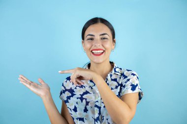 İzole edilmiş mavi arka planda duran genç ve güzel bir kadın gülümsüyor ve elinde bir şey gösteriyor. Boşluğu Kopyala