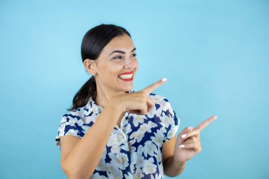 İzole edilmiş mavi arka planda duran genç ve güzel bir kadın şaşırmış ve iki eliyle ve parmaklarıyla işaret ediyor..