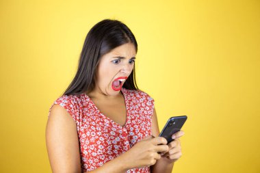 Soyutlanmış sarı arka planda sohbet eden ve telefonuyla şaşıran genç ve güzel bir kadın.