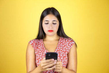 İzole edilmiş sarı arka planda genç ve güzel bir kadın telefonuyla mesajlaşıyor.