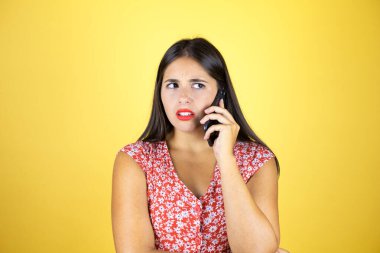 Soyutlanmış sarı arka planda genç ve güzel bir kadın telefonda şaşkın bir ifadeyle konuşuyor.