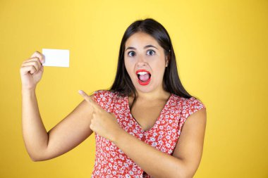 Soyutlanmış sarı arka planda genç ve güzel bir kadın şaşırdı ve beyaz kart gösterdi.