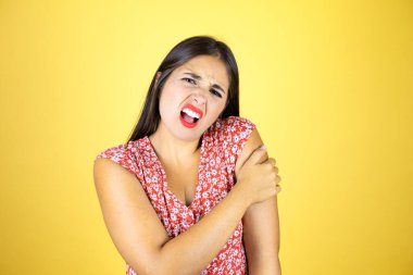 Omzunda ağrı ve acı dolu bir ifadeyle izole edilmiş sarı arka planı olan genç ve güzel bir kadın.