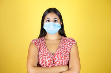 Maskeli ve kolları çaprazlanmış, izole edilmiş sarı arka planı olan genç ve güzel bir kadın. Yüzünde olumsuz ve ciddi bir jest olan uyarı ifadesi.