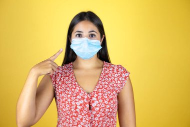 Maskeyi gösteren, izole edilmiş sarı arka planı olan genç ve güzel bir kadın. Yüzünde olumsuz ve ciddi bir jest olan uyarı ifadesi.