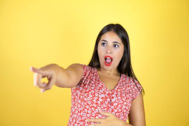 İzole edilmiş sarı arka plan üzerinde genç ve güzel bir kadın. Parmağı önde, ağzı açık, şaşırmış bir ifade. Ön tarafta bir şey var.