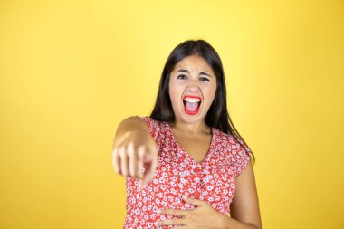 İzole edilmiş sarı arka planda genç ve güzel bir kadın sana gülüyor, eli vücudunun üzerinde kamerayı işaret ediyor, utanç ifadesi.