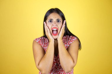 Sarı arka planda yaşayan genç ve güzel bir kadın çığlık atıyor ve elleri ağzında şaşırıyor.
