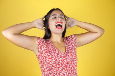 Genç ve güzel bir kadın izole edilmiş sarı arka planda rahatlıyor ve esniyor, kolları ve elleri başının arkasında mutlu bir şekilde gülümsüyor.