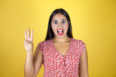 İzole edilmiş sarı arka planda genç ve güzel bir kadın üç numaralı parmaklarıyla işaret ederken kendinden emin ve mutlu bir şekilde gülümsüyor.