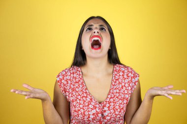 Genç ve güzel bir kadın, izole edilmiş sarı arka planda çılgın ve kızgın bir şekilde bağırıyor ve agresif bir ifade ve kolla bağırıyor.