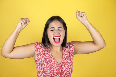 Genç ve güzel bir kadın, izole edilmiş sarı arka planda çok mutlu ve heyecanlı. Kollarını kaldırıp, gülümseyerek ve başarı için çığlık atarak kazanan bir jest yapıyor..