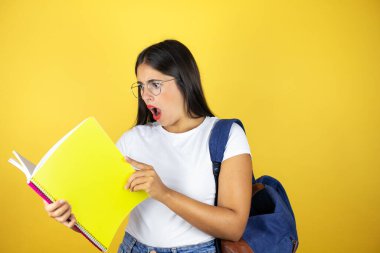 Sırt çantasıyla gezen genç ve güzel bir bayan izole edilmiş sarı arka planda not defteri tutuyor.