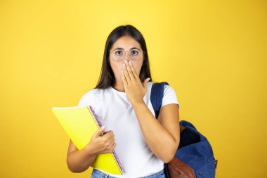 Sırt çantasıyla gezen genç ve güzel bir bayan izole edilmiş sarı arka planda not defteri tutuyor. Ağzını yanlışlıkla ellerle kapatıyor. Gizli konsept.