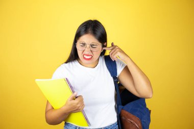 Sırt çantasıyla gezen genç ve güzel bir bayan yüksek sesli müzik sesi için sarı arka plan üzerinde not defteri ile kulakları parmaklarıyla kapatıyor. Sağır kavramı.