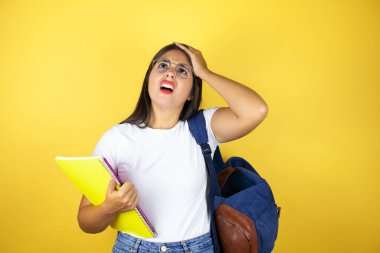Sırt çantasıyla gezen genç ve güzel bir kız. Sarı arka planda defter tutuyor. Bir elini başına koyup sanki bir şey unutmuş gibi gülümsüyor.