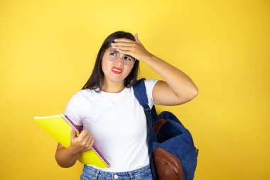 Sırt çantasıyla gezen genç ve güzel bir bayan izole edilmiş sarı arka planda defter tutuyor. Hastalık, ateş, grip ve virüs için alnına dokunuyor.