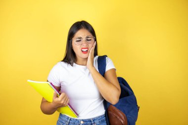 Sırt çantasıyla gezen genç ve güzel bir bayan diş ağrısı ya da diş ağrısı yüzünden ağzı ağrıyan sarı arka plan üzerinde defter taşıyor.
