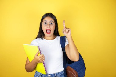 Sırt çantası takan genç ve güzel bir bayan izole edilmiş sarı arka planda defter tutuyor.