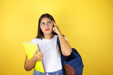 Sırt çantasıyla gezen genç ve güzel bir bayan izole edilmiş sarı arka planda defter tutuyor. Gülümsüyor ve parmağıyla bir fikri olduğunu düşünüyor..