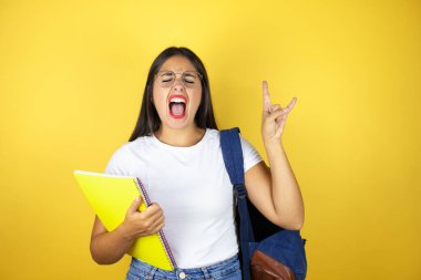 Sırt çantası taşıyan genç ve güzel bir bayan izole edilmiş sarı arka planda defter tutuyor çılgın bir ifadeyle elleri havada rock sembolü yapıyor.