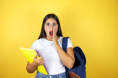 Sırt çantasıyla gezen genç ve güzel bir kız. Elinde not defteri var. Sarı arka planda. Elleri ağzında ve şaşırmış.