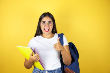 Sırt çantası taşıyan genç ve güzel bir bayan izole edilmiş sarı arka planda defter tutuyor gülümsüyor ve baş parmağıyla işaret veriyor.