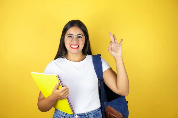 Young beautiful student woman wearing backpack holding notebook over isolated yellow background doing ok sign with fingers and smiling, excellent symbol