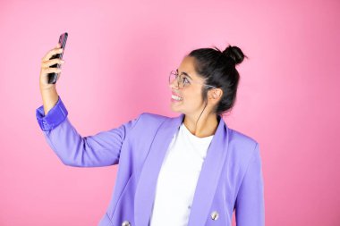 Genç ve güzel bir iş kadını pembe arka planda telefonuyla selfie çekiyor.
