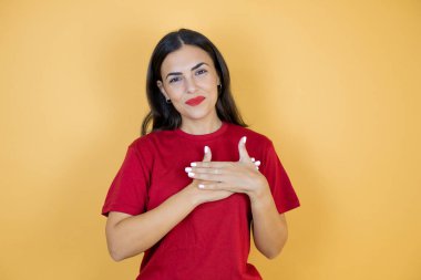 Güzel bir kadın, izole edilmiş sarı arka planda elleri göğsünde ve yüzünde minnet dolu bir jest ile gülümsüyor..