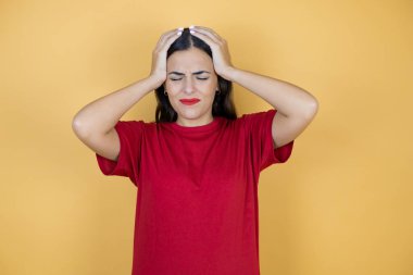 Genç ve güzel bir kadın, sarı arka planda, baş ağrısı, çaresiz ve stresli, çünkü elleri başının üstünde acı ve migren var..