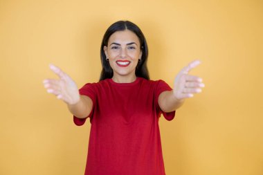 SarıImak için kollarını açmış kameraya gülümseyen, izole edilmiş sarı arka planda genç ve güzel bir kadın..