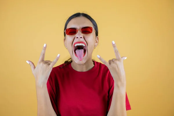 İzole edilmiş sarı arka planda kırmızı güneş gözlüğü takan genç ve güzel bir kadın çılgın bir ifadeyle el kaldırarak rock sembolü yapıyor..