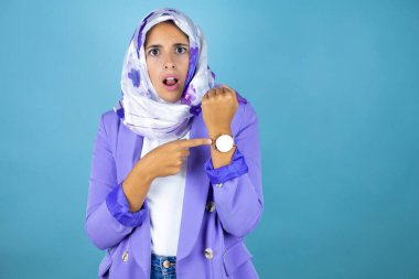 Genç, güzel bir Arap kadın, tecrit edilmiş mavi arka plan üzerinde İslami başörtüsü takıyor. Geç olduğu için şaşırıp saatini işaret ediyor.