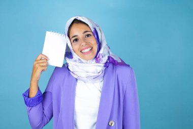 Genç, güzel bir Arap kadın, izole edilmiş mavi arka plan üzerine tesettür takıyor. Elinde boş bir defter var ve gülümsüyor.
