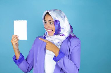 Genç, güzel, Arap bir kadın, izole edilmiş mavi arka planda tesettür takıyor, gülümsüyor ve elindeki boş defteri işaret ediyor.