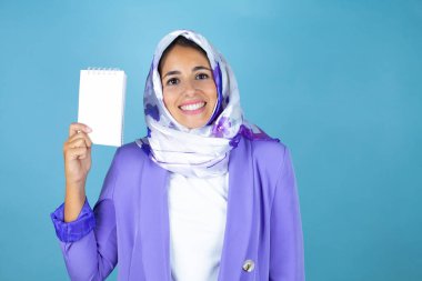 Genç, güzel bir Arap kadın, izole edilmiş mavi arka plan üzerine tesettür takıyor. Elinde boş bir defter var ve gülümsüyor.