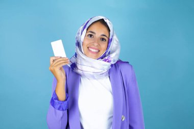 Genç, güzel bir Arap kadın, izole edilmiş mavi arka plan üzerinde İslami başörtüsü takıyor gülümsüyor ve elinde beyaz bir kart tutuyor.