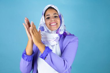 Genç, güzel bir Arap kadın, izole edilmiş mavi arka planda, tesettürlü, tesettürlü, mutlu ve neşeli elleri alkışlayan, İslami başörtüsü takıyor.