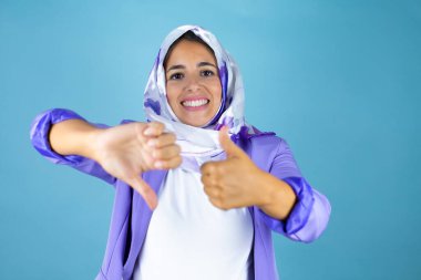Genç, güzel bir Arap kadın, izole edilmiş mavi arka plan üzerinde tesettüre girmiş, başparmak kaldırıyor, anlaşmazlık ve uzlaşma ifadesi sergiliyor. Çılgın çatışma