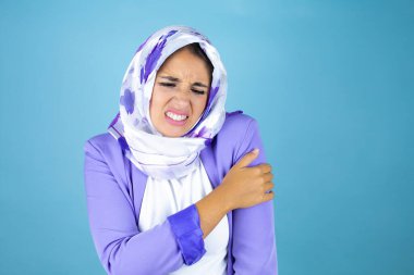 Genç, güzel bir Arap kadın, izole edilmiş mavi arka plan üzerinde İslami başörtüsü takıyor. Omzunda ağrı ve acı dolu bir ifade var.