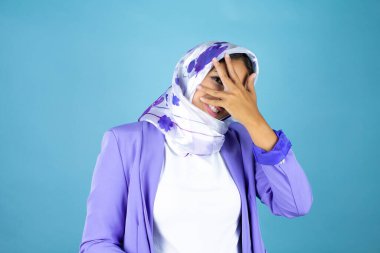 Genç, güzel bir Arap kadın, izole edilmiş mavi arka plan üzerine tesettür takmış. Şok olmuş bir şekilde yüzünü ve gözlerini kapamış, utanmış bir ifadeyle parmaklarının arasından bakıyor.