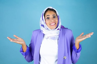 Genç, güzel bir Arap kadın, izole edilmiş mavi arka plan üzerine tesettür takıyor. Kolları ve elleri havada, kafası karışmış bir ifade.