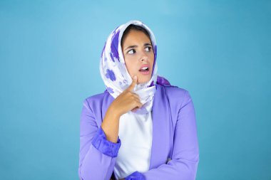Genç, güzel bir Arap kadın, izole edilmiş mavi arka planda İslami başörtüsü takıyor. Parmağıyla çenesinde bir sorusu olduğunu düşünüyor.