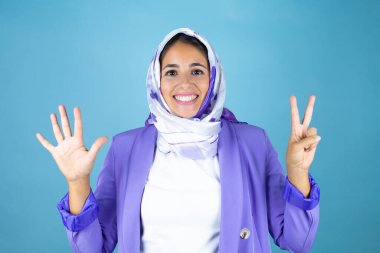 Genç, güzel bir Arap kadın, izole edilmiş mavi arka planda İslami başörtüsü takıyor ve yedinci parmaklarıyla işaret ederken kendinden emin ve mutlu bir şekilde gülümsüyor.