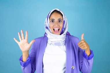 Genç, güzel bir Arap kadın, izole edilmiş mavi arka planda İslami başörtüsü takıyor ve altı numaralı parmaklarıyla işaret ederken kendinden emin ve mutlu bir şekilde gülümsüyor.