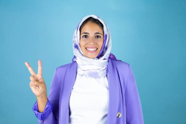 Genç, güzel bir Arap kadın, izole edilmiş mavi arka planda İslami başörtüsü takıyor ve iki numaralı parmaklarıyla işaret ederken kendinden emin ve mutlu bir şekilde gülümsüyor.