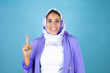 Genç, güzel bir Arap kadın, izole edilmiş mavi arka planda İslami başörtüsü takıyor ve parmaklarıyla bir numarayı gösterirken kendinden emin ve mutlu bir şekilde gülümsüyor.