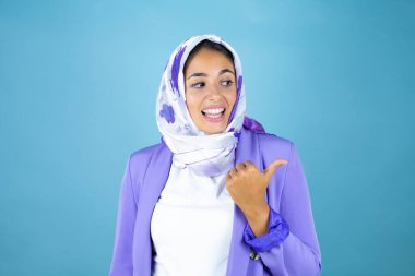 Genç, güzel bir Arap kadın, izole edilmiş mavi arka planda başparmağıyla gülümseyerek ve işaret ederek başörtüsü takıyor.