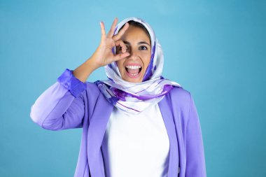 Genç, güzel bir Arap kadın, izole edilmiş mavi arka planda İslami başörtüsü takıyor. Tamam, gülümseyen yüzle, gözleriyle parmaklarından bakıyor.
